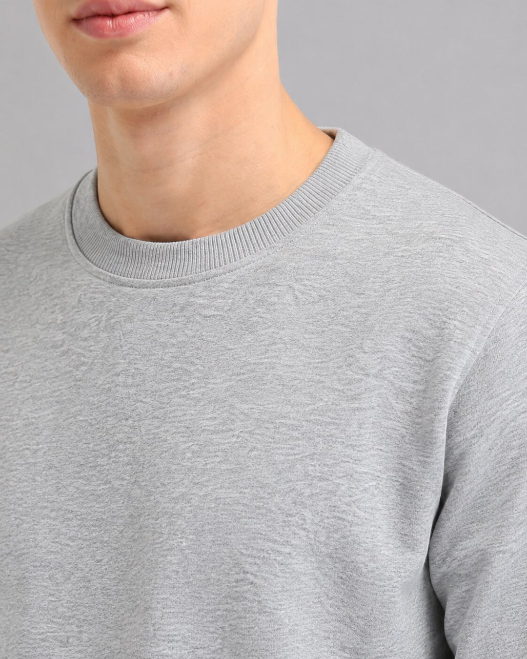 Close-up of a person wearing a light gray crewneck sweatshirt, showing the ribbed collar and textured fabric against a plain gray background.