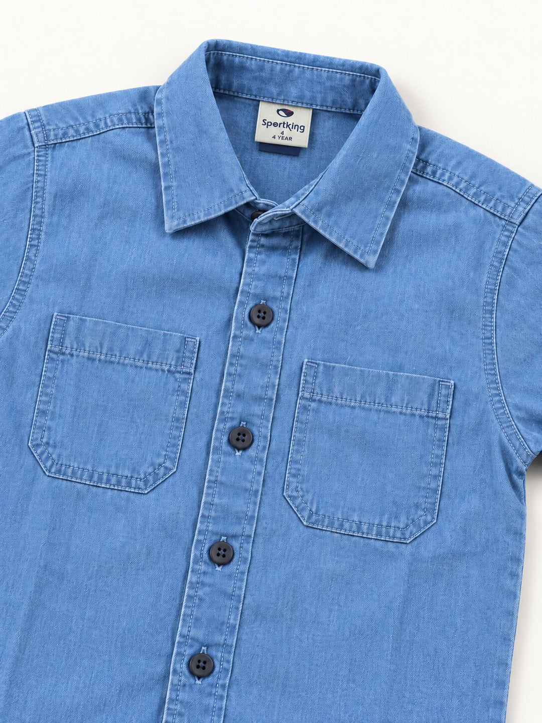 A close-up of a light blue denim button-down shirt with two chest pockets and dark buttons, laid flat against a white background.