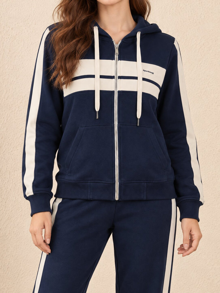 A woman wearing a navy blue zip-up hoodie with cream-colored side stripes, a horizontal chest stripe, and matching navy sweatpants with cream side stripes, set against a textured beige wall.