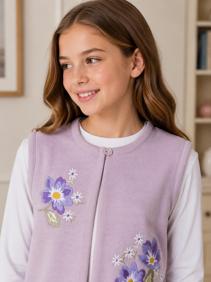 A young girl with long brown hair smiling and looking to the side, wearing a lavender zippered fleece vest with embroidered purple and white flowers over a white long-sleeve shirt.