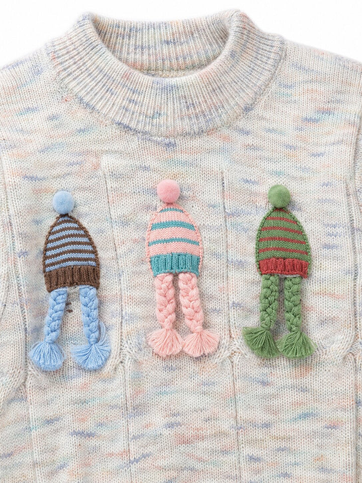 Close-up of a knit sweater with three colorful crocheted hat appliqués on the chest, each with braided tassels. The sweater has a marled cream base with subtle pastel blue and pink flecks.