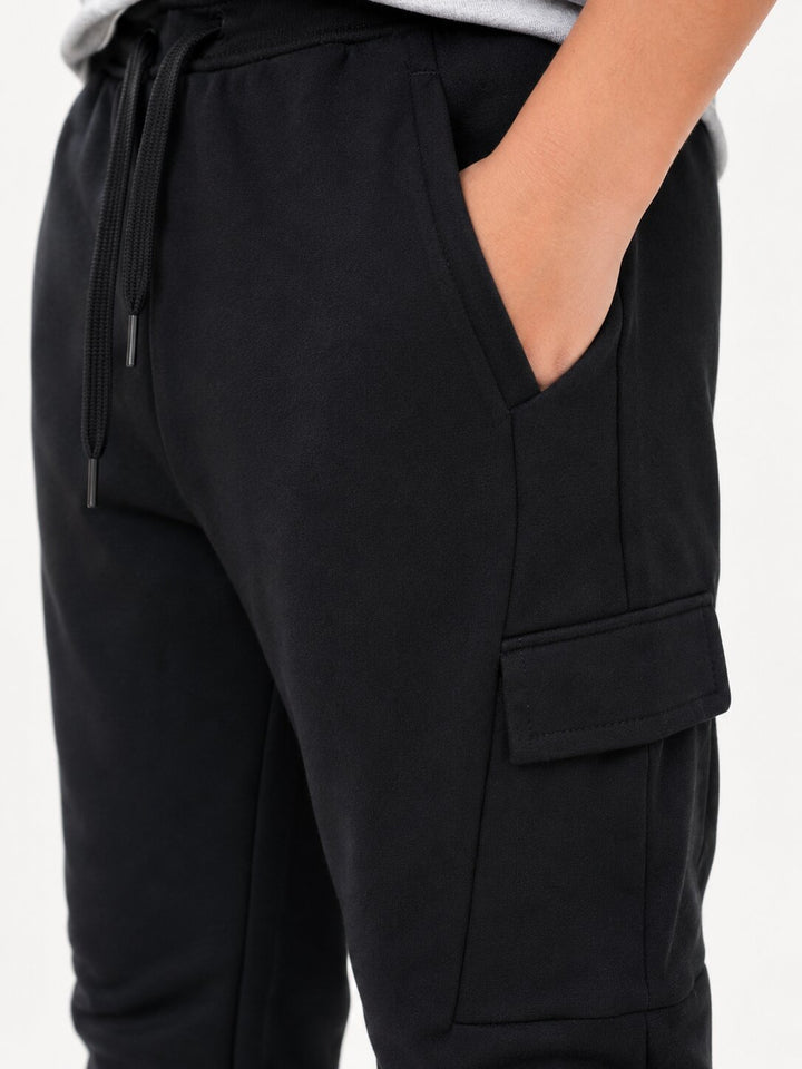 Close-up of a person wearing black jogger-style cargo pants with a drawstring waist, side pocket, and flap cargo pocket, against a plain light background.