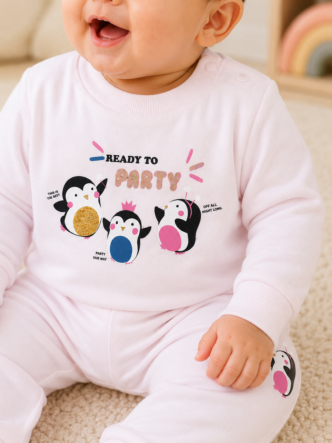 A baby wearing a light pink long-sleeve romper with a penguin-themed graphic that reads 'READY TO PARTY' on the chest, sitting on a beige carpeted floor.