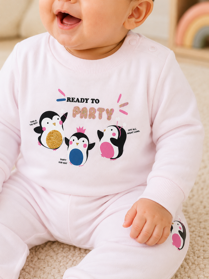 A baby wearing a light pink long-sleeve romper with a penguin-themed graphic that reads 'READY TO PARTY' on the chest, sitting on a beige carpeted floor.