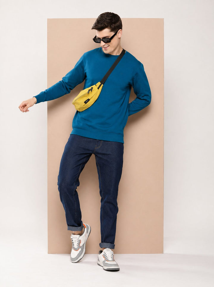 A man wearing a blue crewneck sweatshirt, dark blue jeans, and white sneakers, accessorized with a bright yellow crossbody fanny pack and black sunglasses, posing in front of a beige rectangular backdrop.