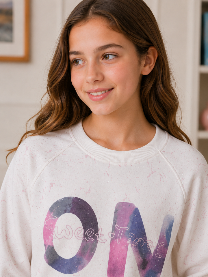 A young girl with long brown hair wearing a white crewneck sweatshirt with a pink and purple watercolor 'ON' graphic, smiling in an indoor setting.