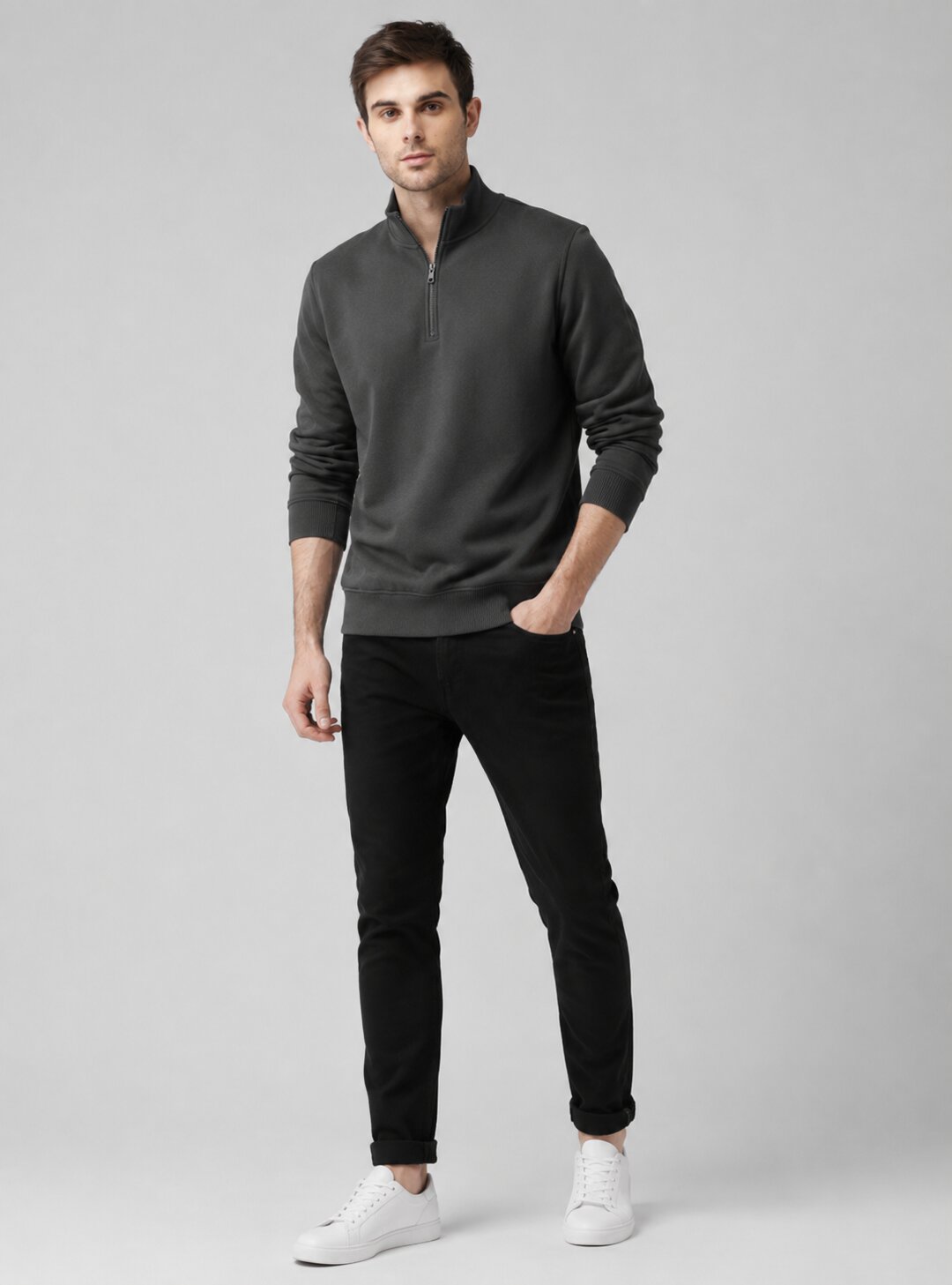 A man modeling a dark gray quarter-zip pullover sweater, black slim-fit jeans, and white sneakers against a solid light gray studio background.