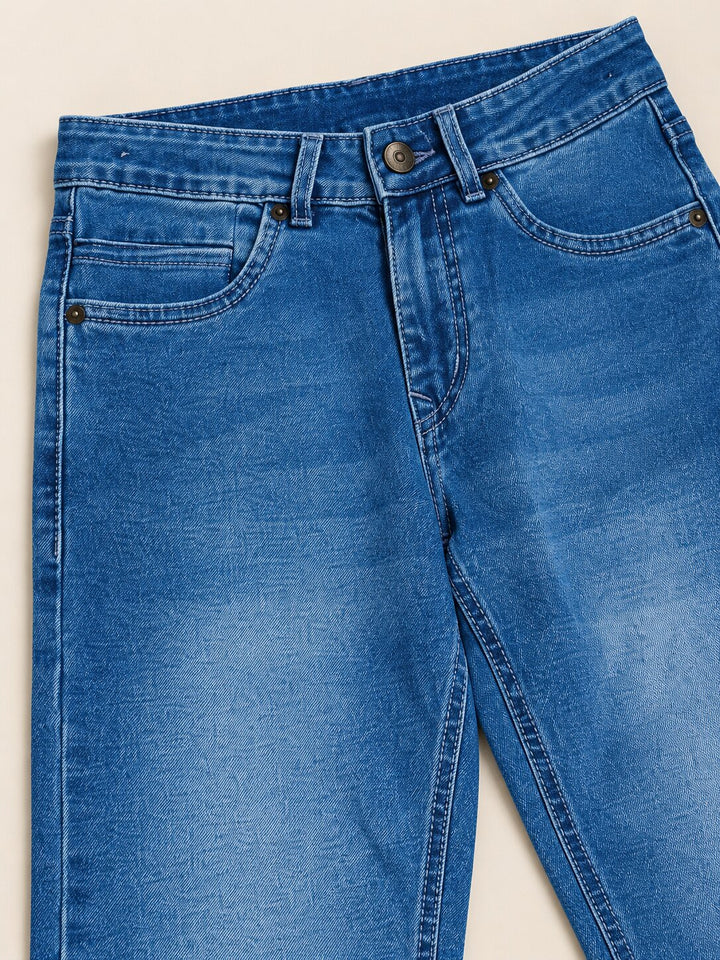 Close-up of blue denim jeans laid flat on a light beige background, showing the waistband, belt loops, front pockets, and leg area.