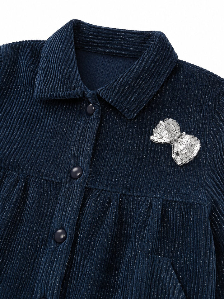 Close-up of a dark navy corduroy button-front garment with a pointed collar and a silver bow-shaped brooch on the chest, shown against a plain white background.