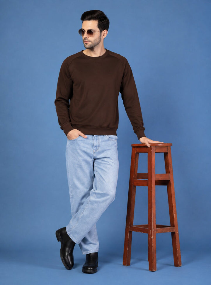 A man wearing a dark brown crewneck sweatshirt, light blue jeans, and black shoes stands next to a wooden stool against a solid blue studio background.