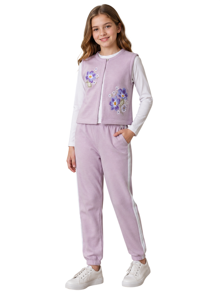 A young girl modeling a lavender two-piece tracksuit featuring a zippered vest with floral embroidery and matching jogger pants with white side stripes, paired with a white long-sleeve shirt and white sneakers.