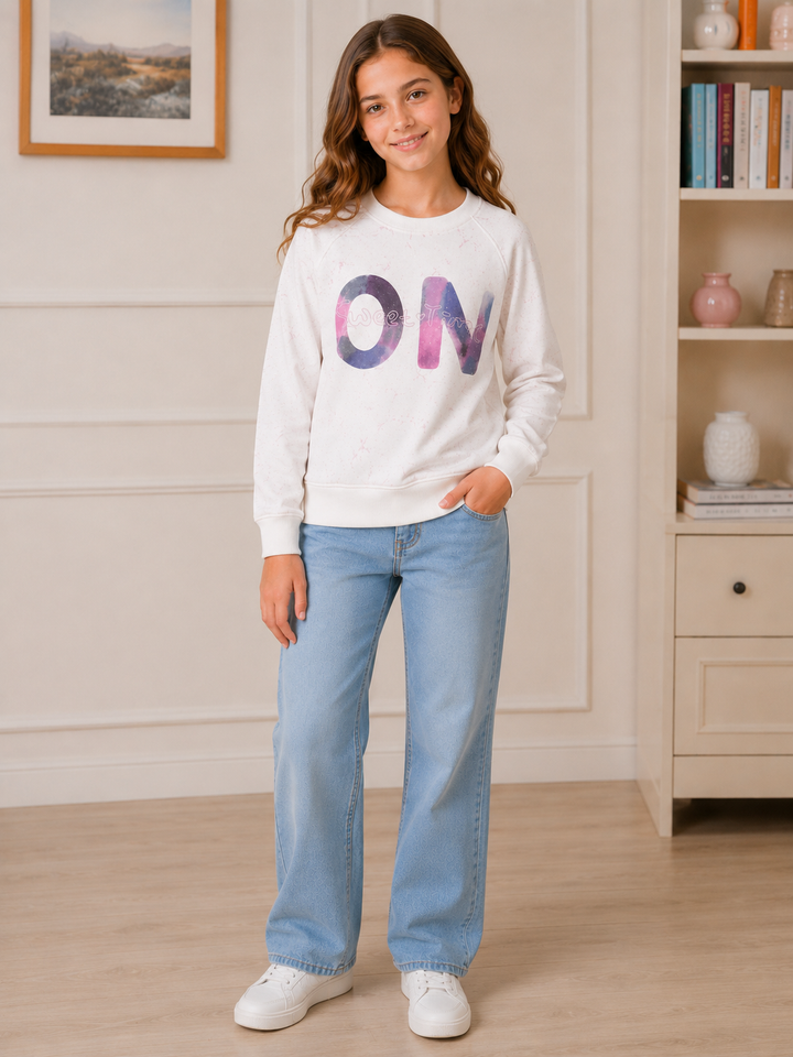 A young girl stands in a bright room wearing a white crewneck sweatshirt with a colorful 'ON' graphic on the front, paired with light blue jeans and white sneakers.