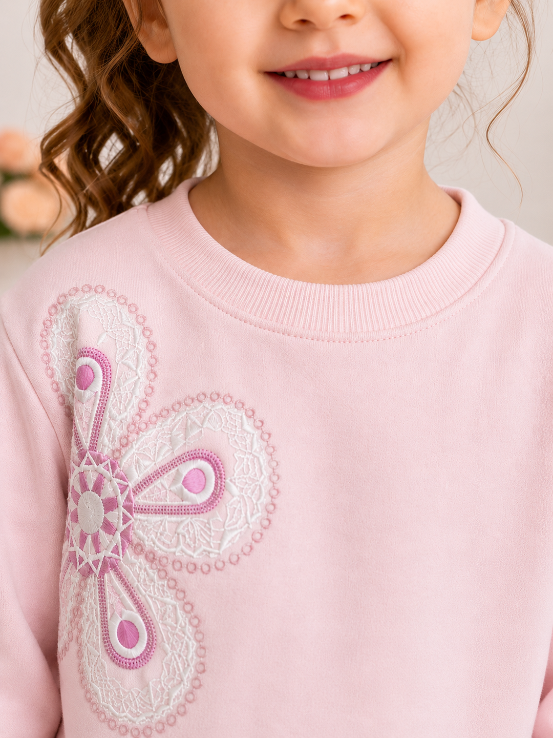 A close-up of a young girl wearing a light pink crewneck sweatshirt with a large white and pink butterfly applique on the front.