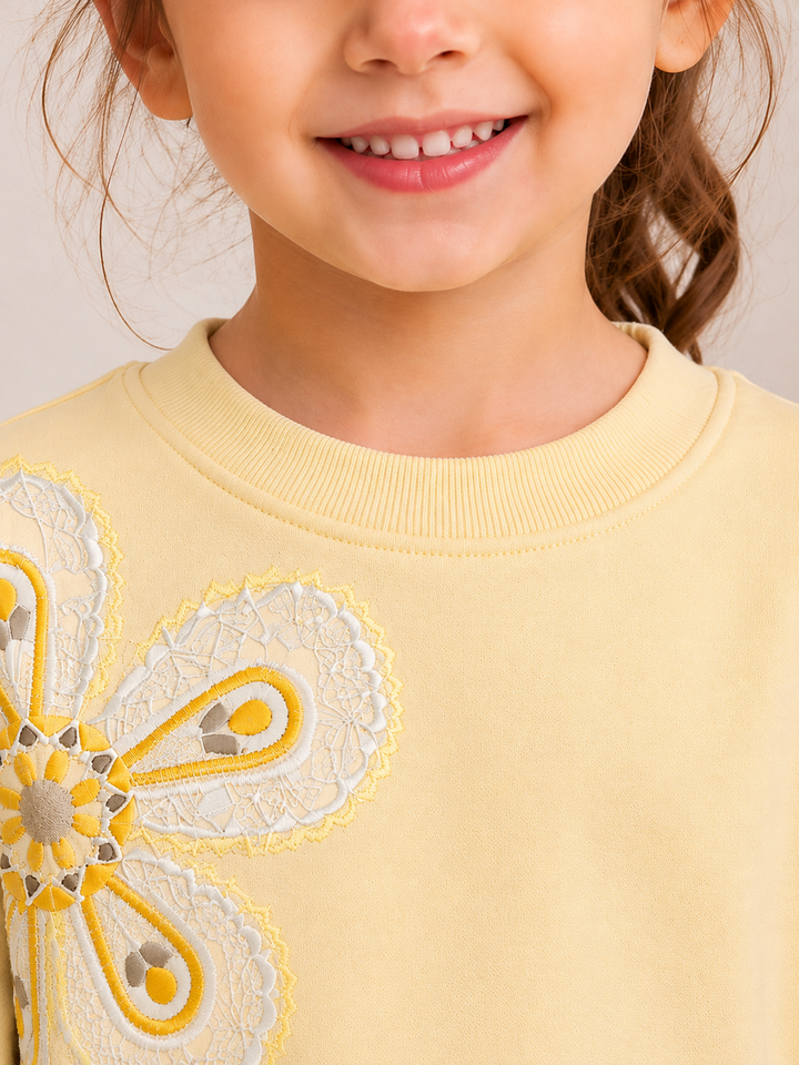 Close-up of a child wearing a pale yellow crewneck sweatshirt with a large white and yellow embroidered floral applique on the chest.