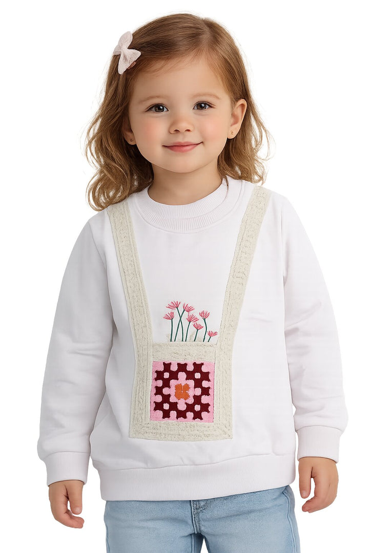 A young girl with light brown hair and a pink bow hair clip is wearing a white crewneck sweatshirt with a decorative front pocket design. The pocket design features pink flowers and a pink and red quilted pattern with beige trim.