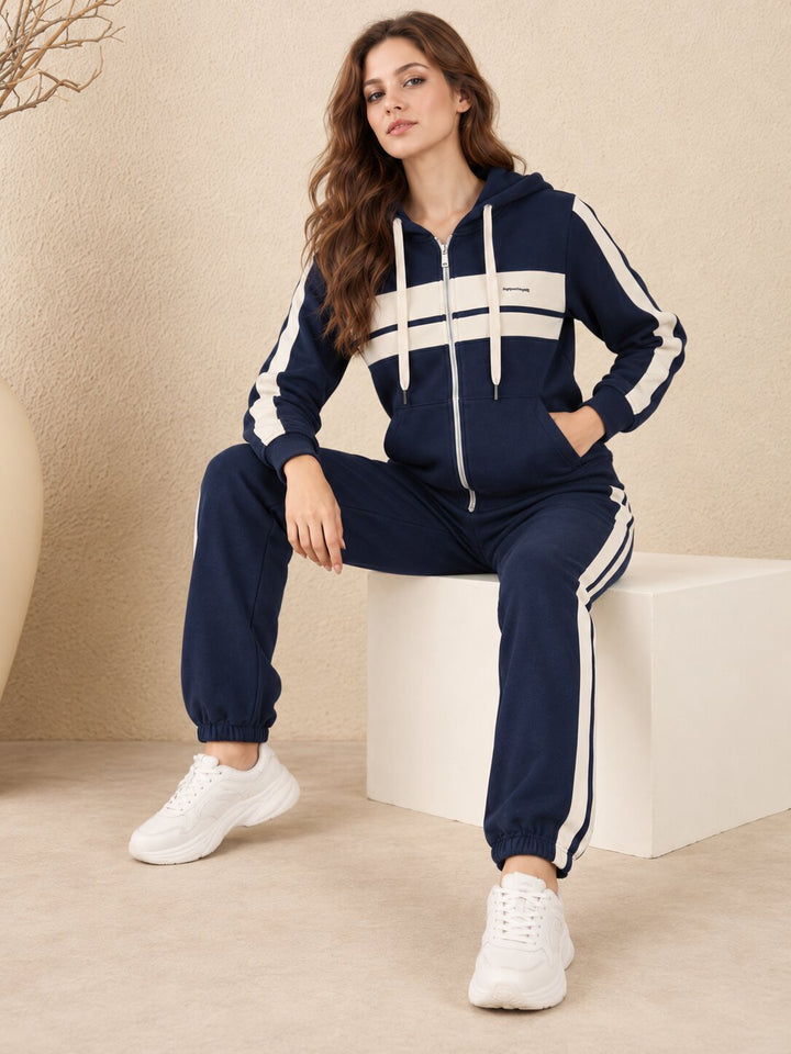 A woman wearing a navy blue and white tracksuit with white sneakers, sitting on a white block in a beige room.