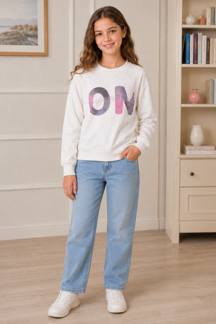 A young girl stands in a bright room wearing a white crewneck sweatshirt with a colorful 'OM' graphic on the front, paired with light blue jeans and white sneakers.