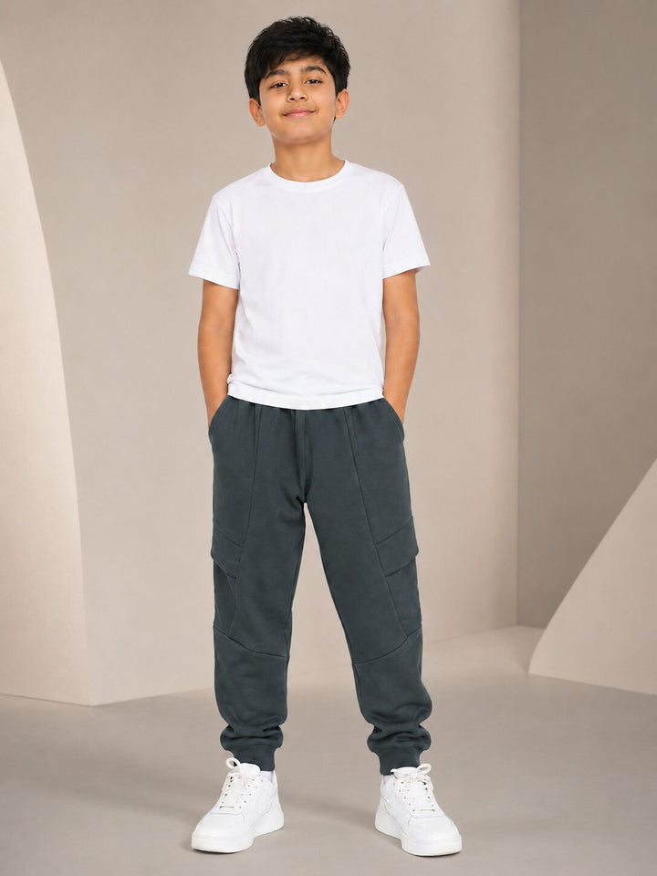 A young boy standing in a studio setting, wearing a plain white crew-neck t-shirt, dark gray jogger-style pants with side cargo pockets, and white sneakers.