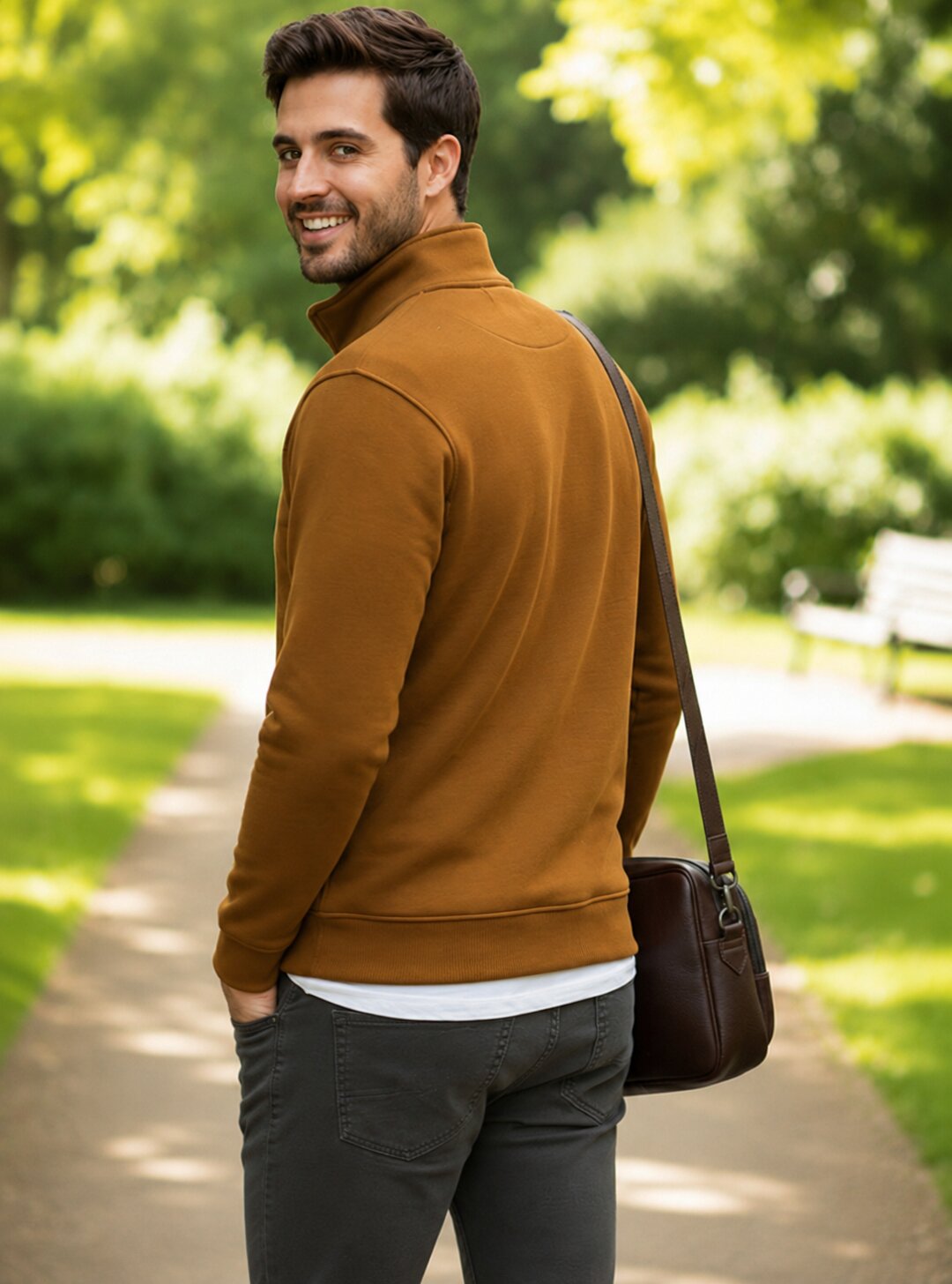 A man wearing a brown mock-neck long-sleeve sweater, dark gray pants, and carrying a brown leather shoulder bag, standing on a park path with green trees in the background.