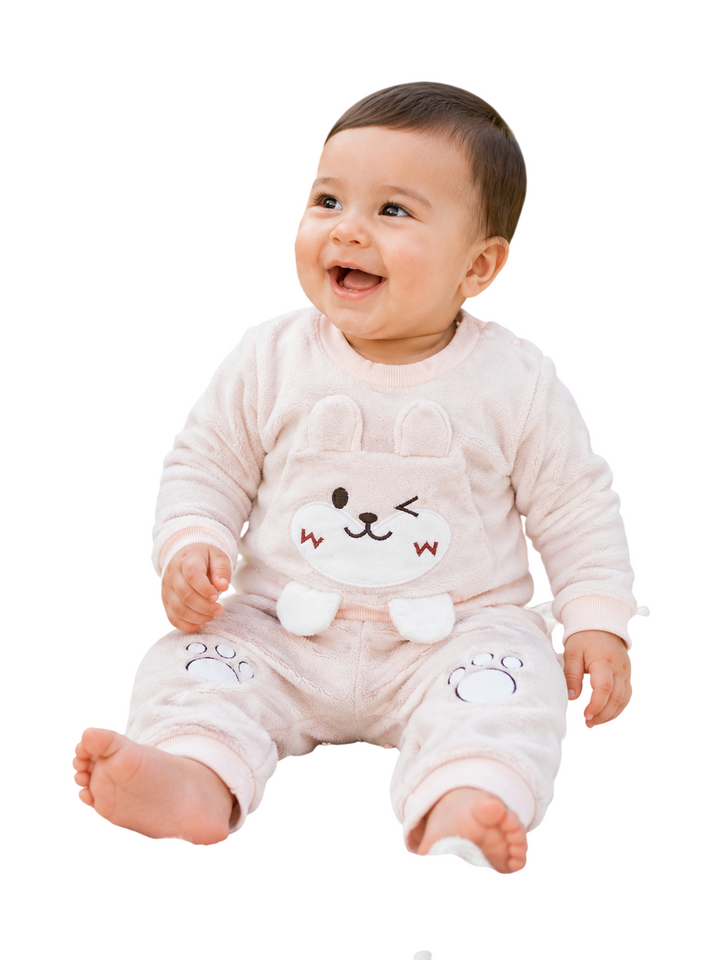 A smiling baby sitting and wearing a light pink plush one-piece pajama with a cartoon bunny face on the front, photographed against a plain white background.