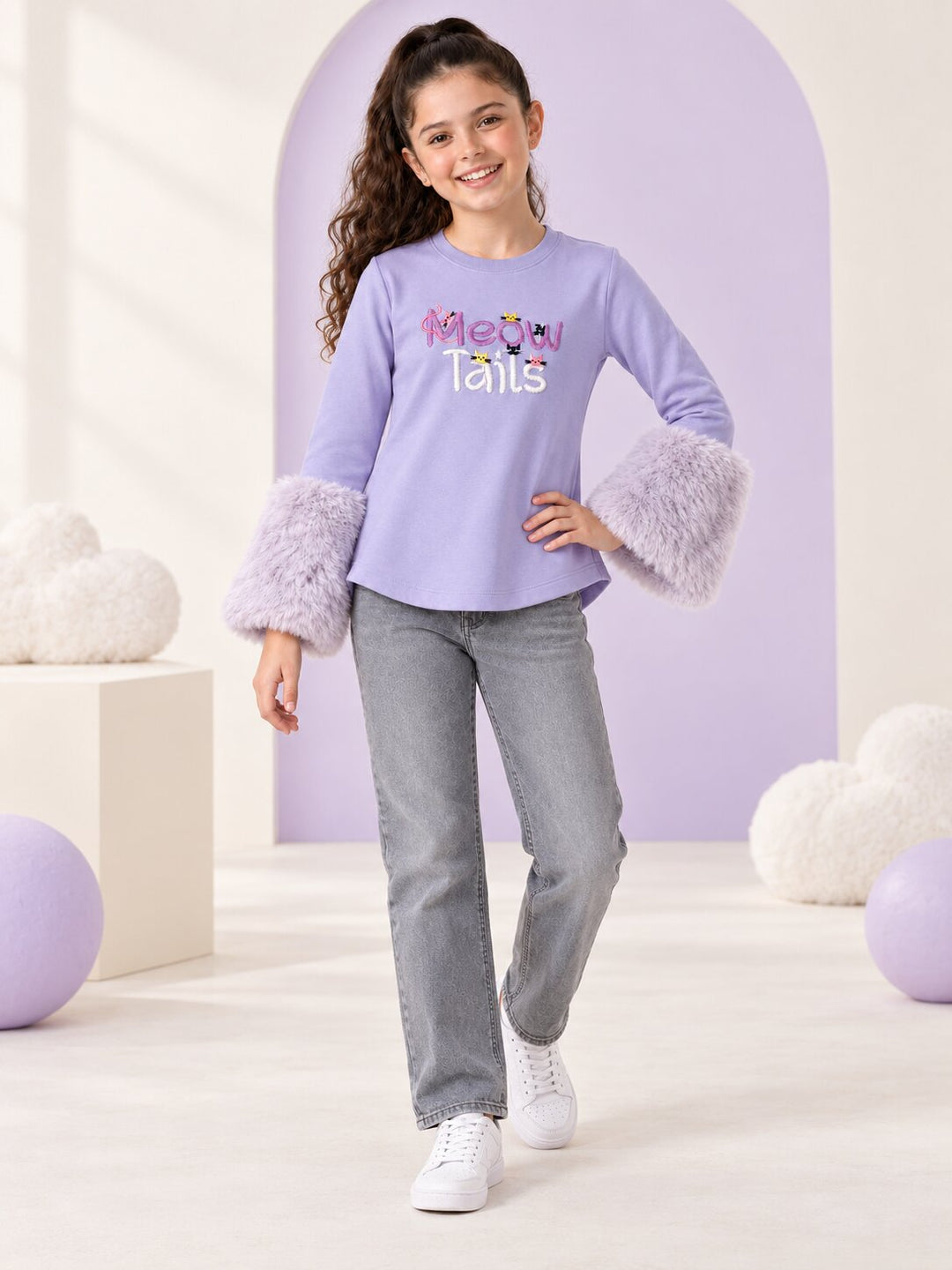A young girl models a lavender long-sleeve t-shirt with fuzzy lavender cuffs and a 'Mom Tarts' graphic on the chest, paired with gray jeans and white sneakers in a pastel studio setting.