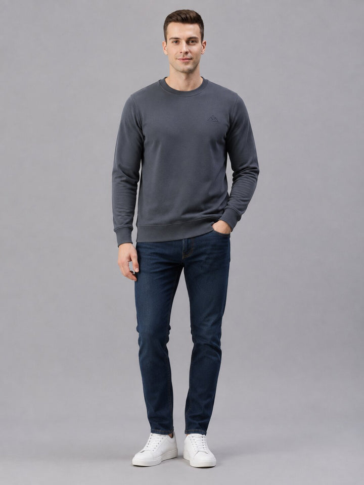 A man modeling a dark gray crewneck sweatshirt, dark blue slim-fit jeans, and white sneakers against a solid gray studio background.