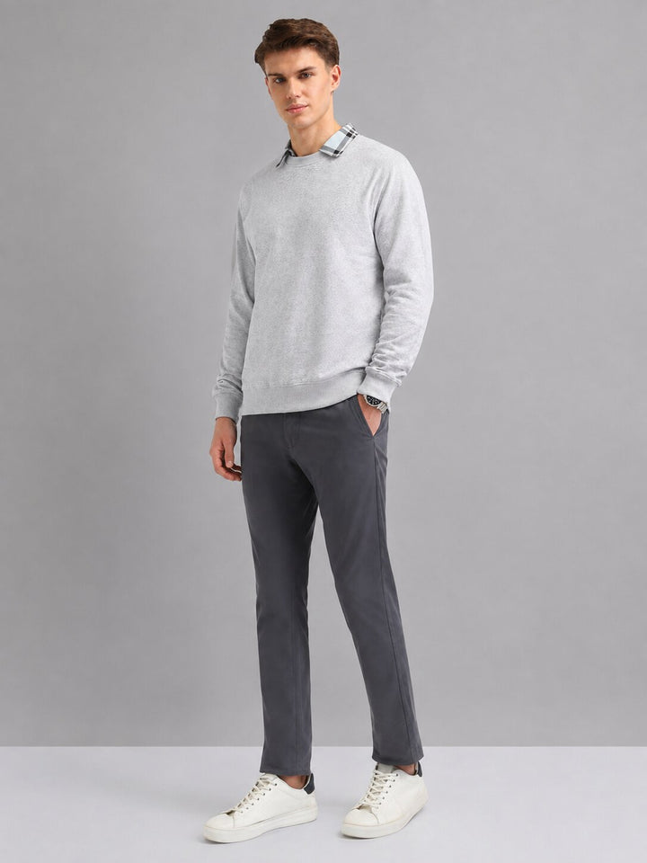 A man modeling a light gray crewneck sweater with a subtle plaid collar detail, paired with dark gray trousers and white sneakers, against a solid gray studio background.
