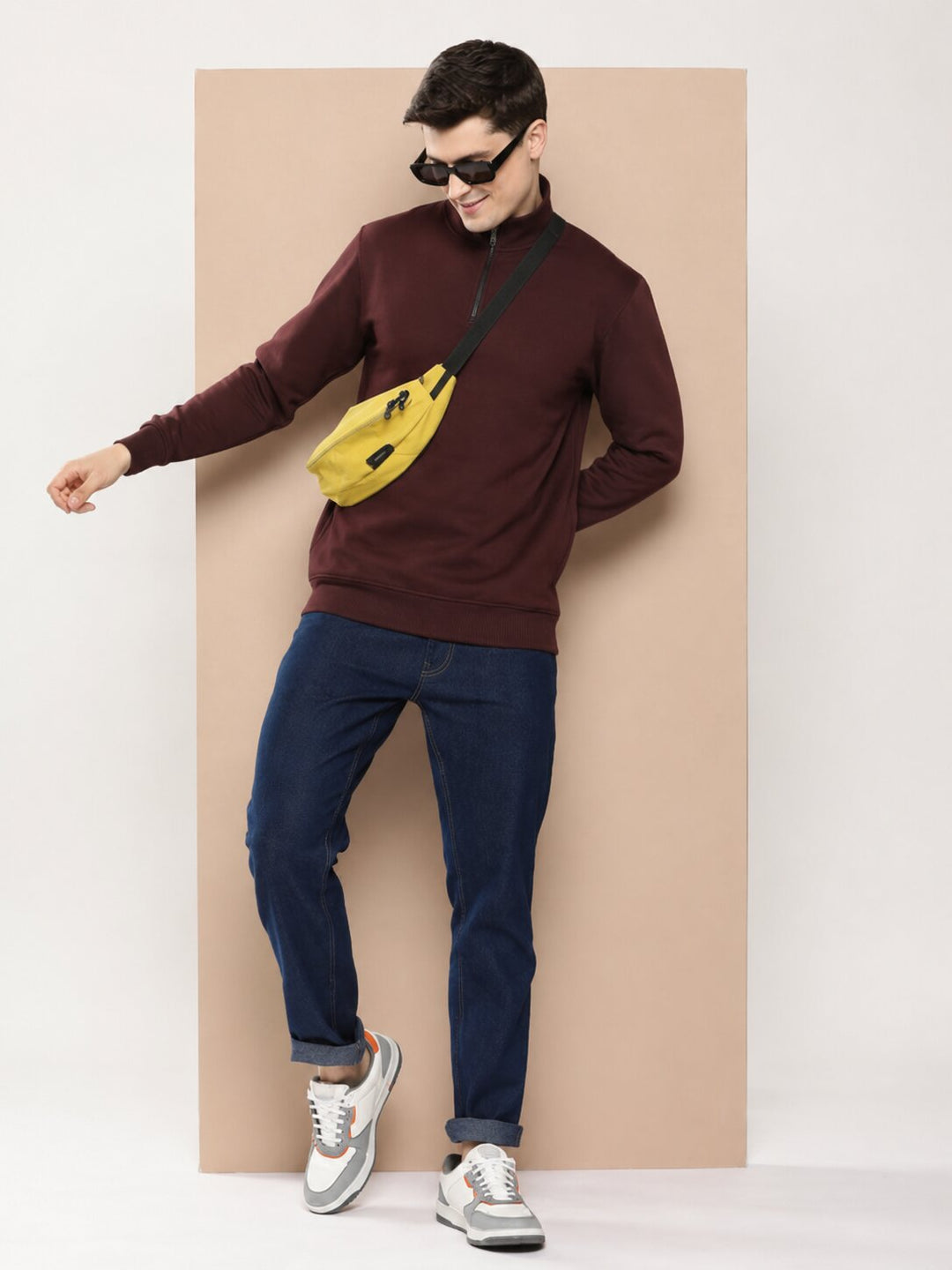 A man modeling a dark burgundy mock-neck sweater, navy blue jeans, and white sneakers, wearing a bright yellow crossbody fanny pack and black sunglasses, posing in front of a beige rectangular backdrop.