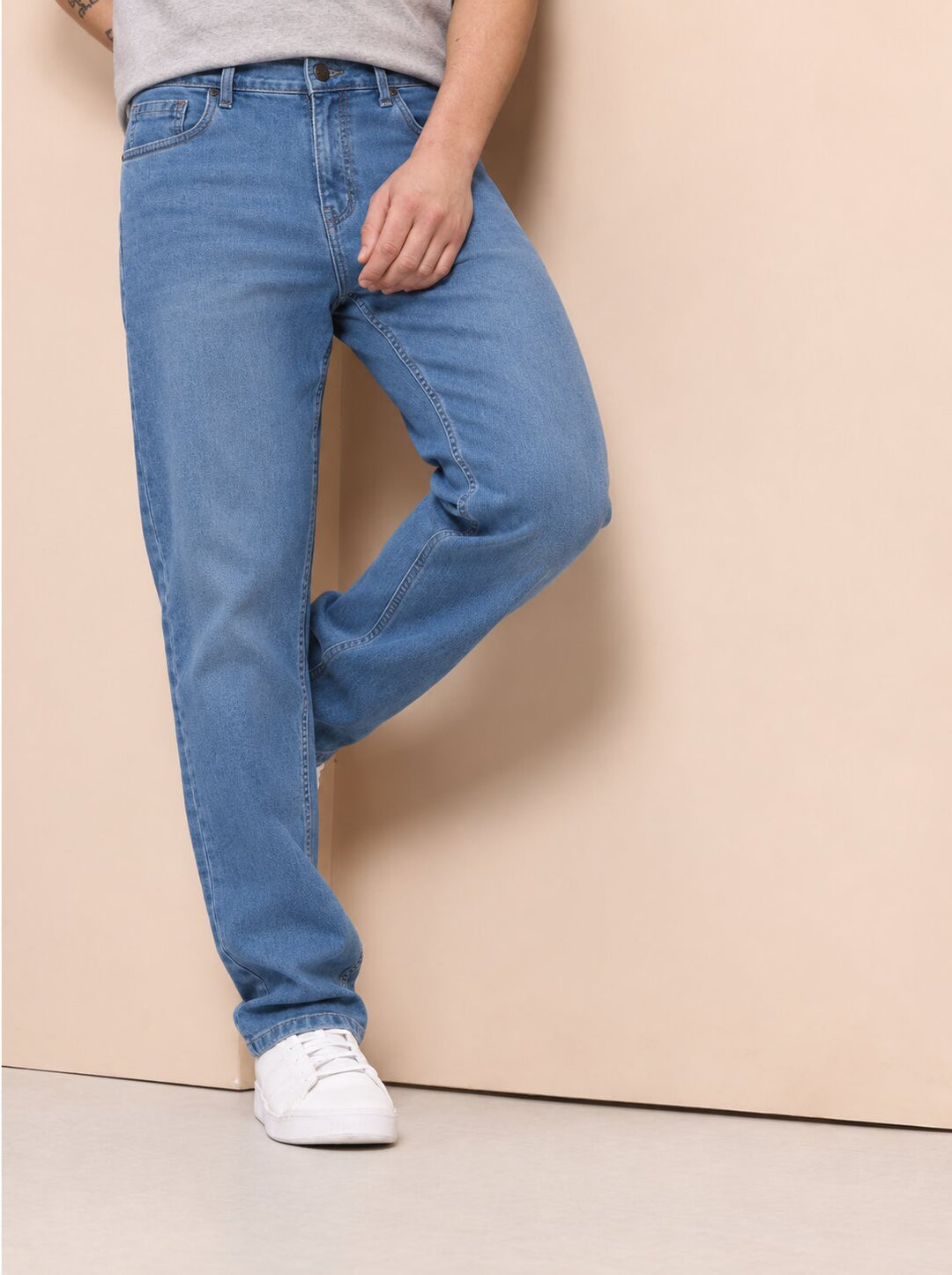 A person wearing light blue straight-leg jeans and white sneakers, standing against a plain beige wall.