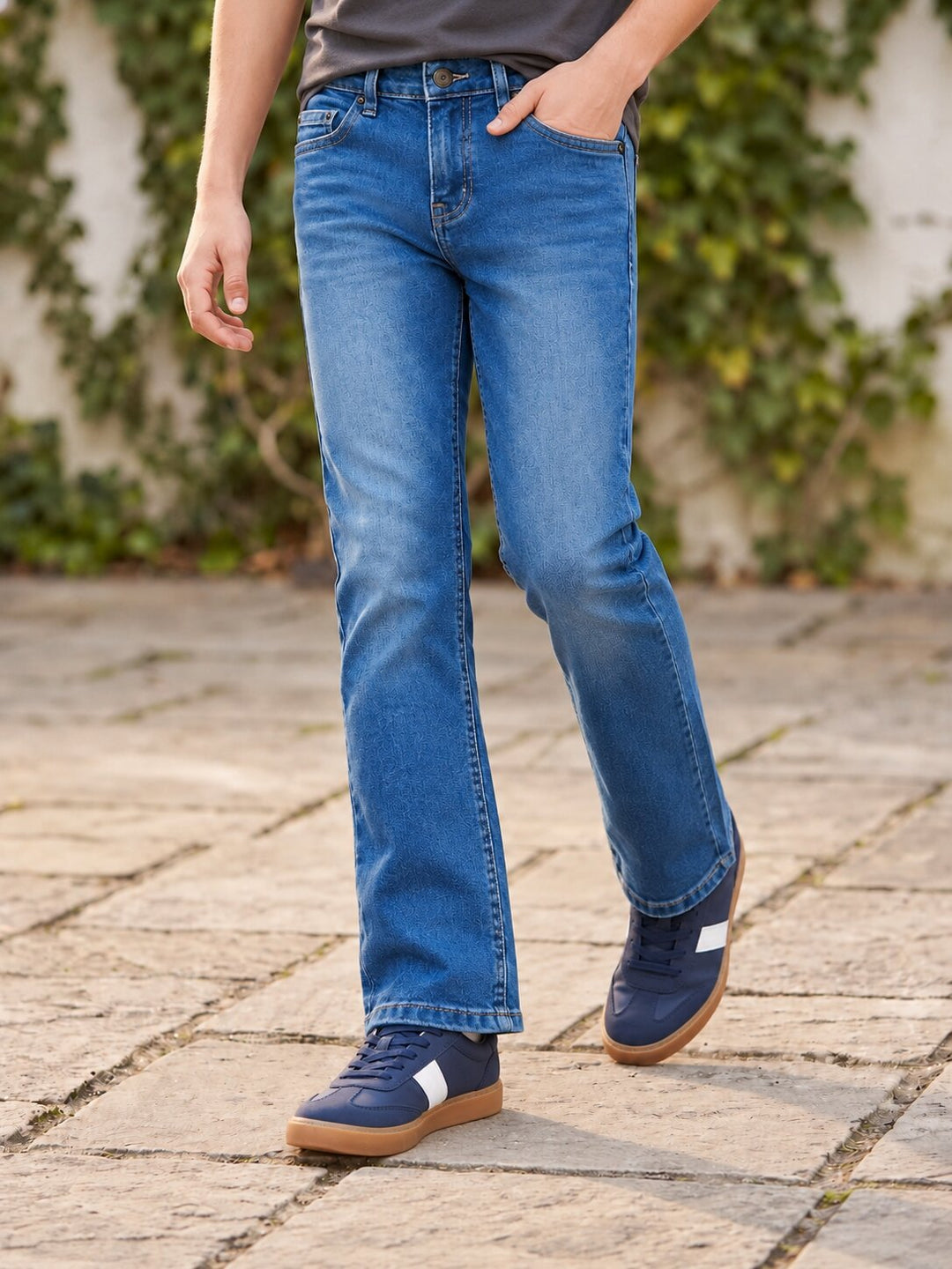 A person wearing blue denim jeans with a straight-to-medium leg, styled with a dark gray t-shirt and navy blue sneakers with white side stripes, standing on a stone paved path with green ivy in the background.