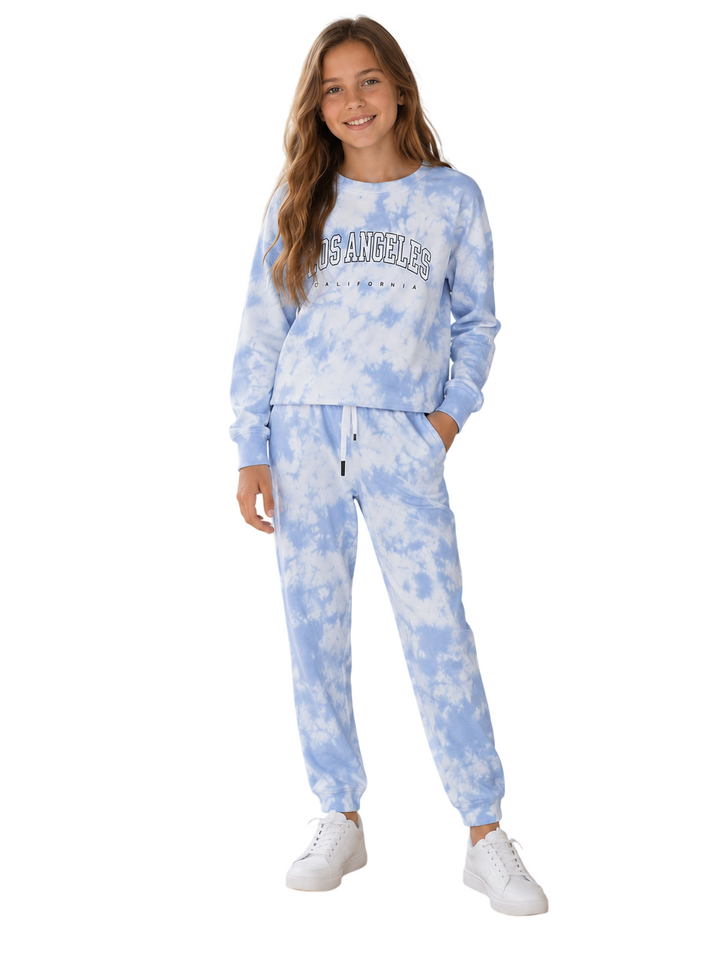A young girl modeling a light blue and white tie-dye sweatshirt and jogger pants set with white sneakers, against a plain white background.