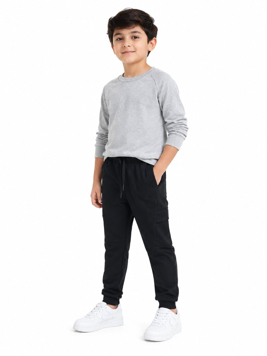 A young boy modeling a light gray long-sleeve raglan sweatshirt and black jogger pants with white sneakers, standing against a solid white background.
