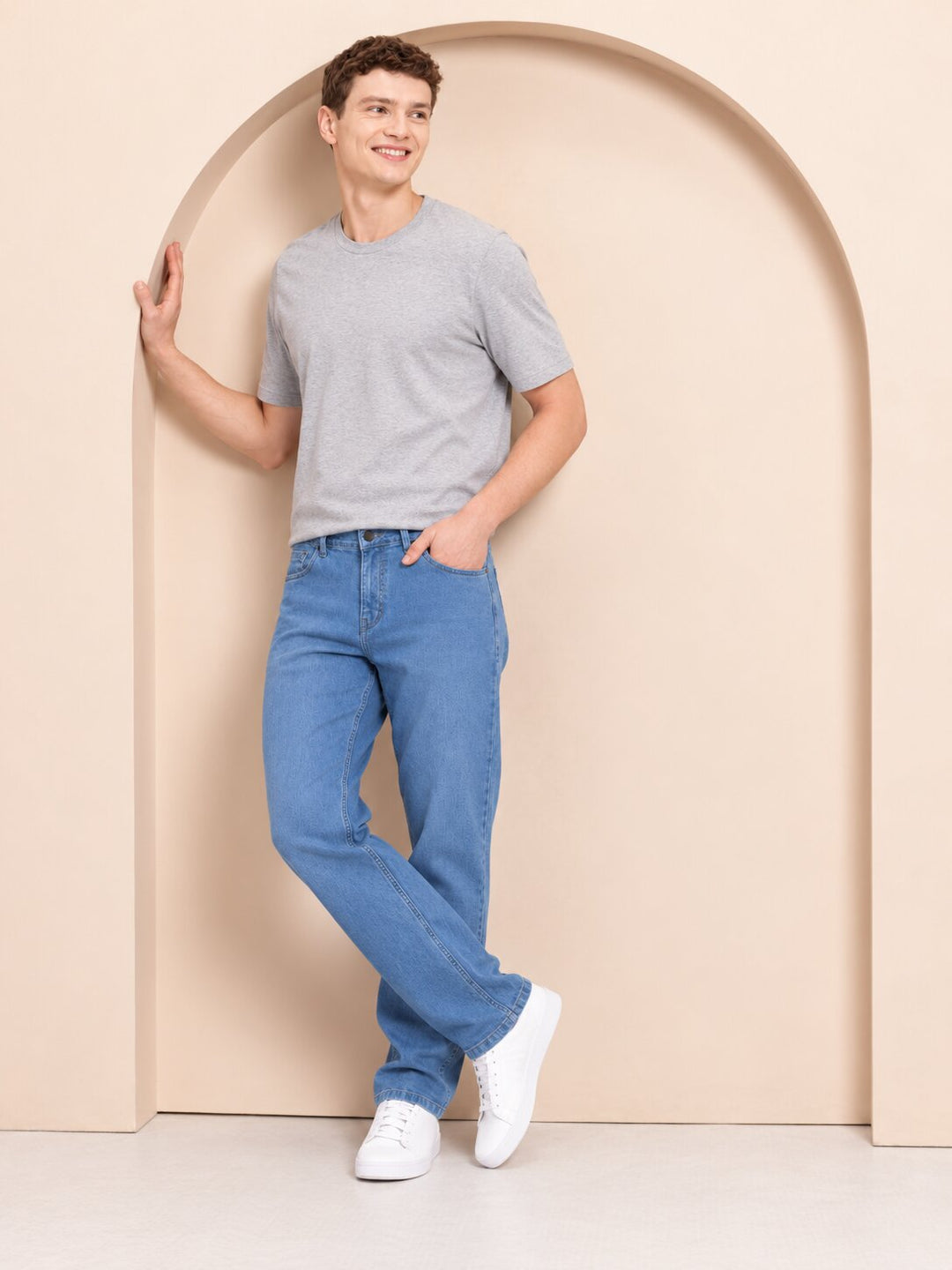 A man wearing a light gray crewneck t-shirt, blue jeans, and white sneakers, posing in front of a beige arched wall.