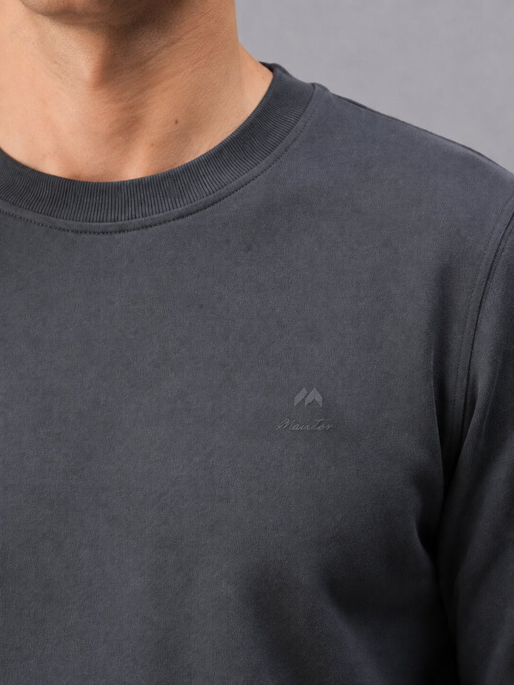 Close-up of a person wearing a dark charcoal gray crewneck sweatshirt with a subtle tonal embroidered logo on the left chest.