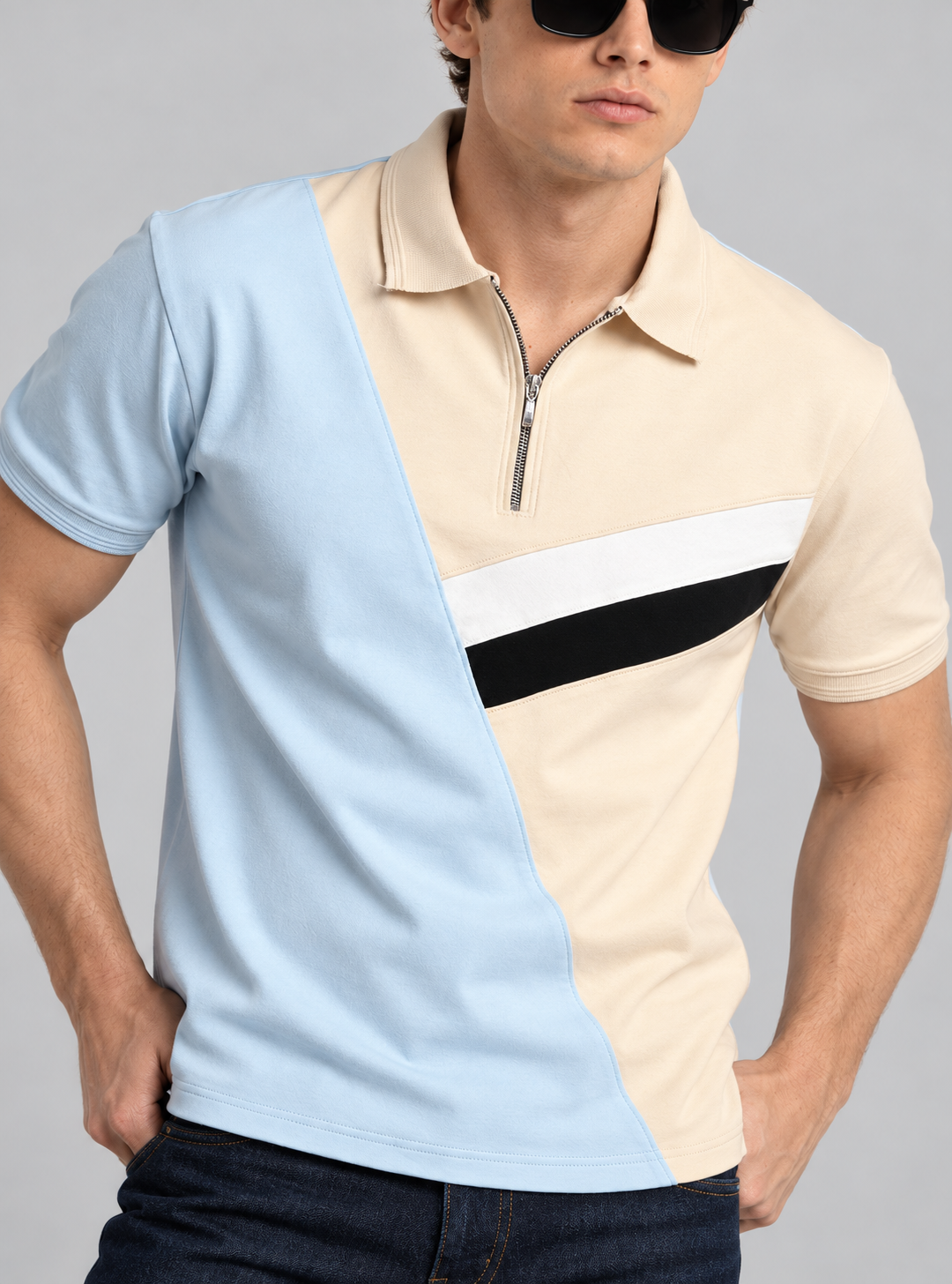 A man wearing a color-blocked polo shirt with a light blue diagonal panel, a cream upper panel, a white horizontal stripe, and a black diagonal stripe, paired with dark sunglasses and dark jeans against a solid gray background.