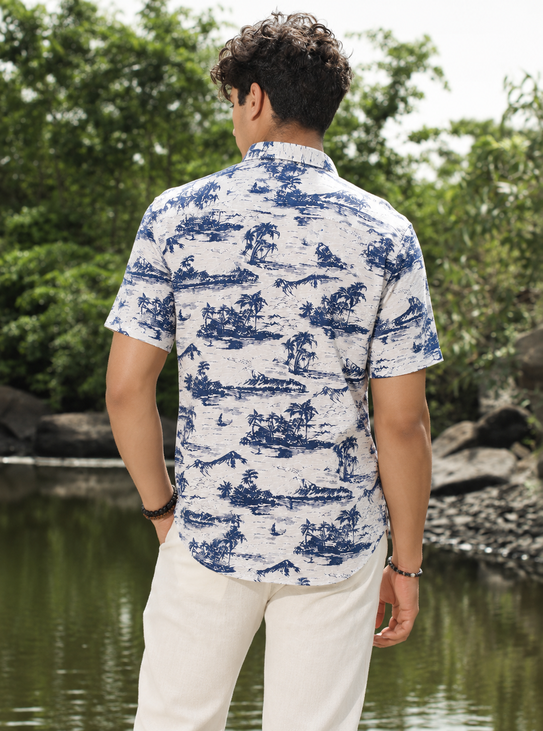A man viewed from behind wearing a short-sleeve button-up shirt with a blue scenic toile de Jouy pattern on a white background, paired with off-white trousers, standing outdoors near a body of water and green trees.