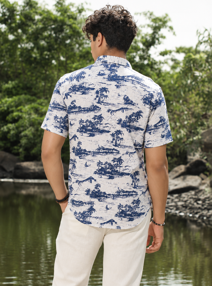 A man viewed from behind wearing a short-sleeve button-up shirt with a blue scenic toile de Jouy pattern on a white background, paired with off-white trousers, standing outdoors near a body of water and green trees.