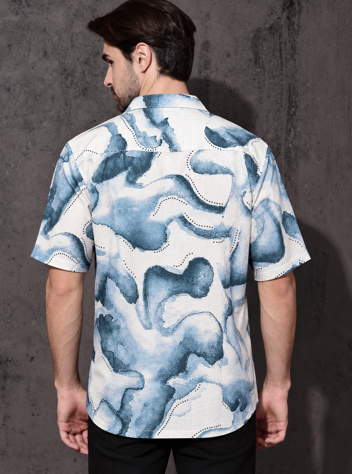 A man viewed from the back wearing a short-sleeve button-up shirt with an abstract blue and white watercolor-style print, paired with black pants, standing against a dark textured wall.