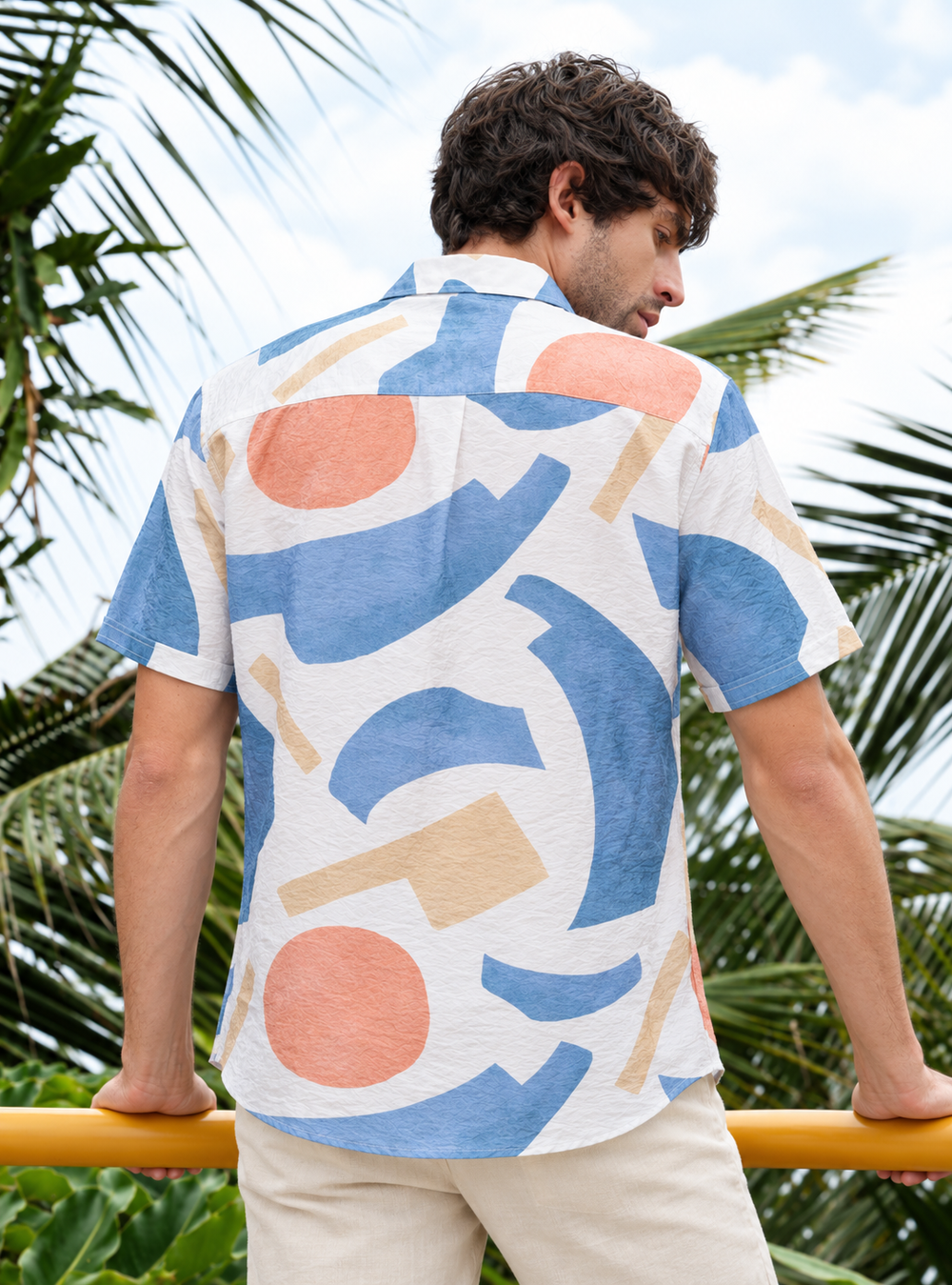 A man wearing a short-sleeve button-up shirt with an abstract geometric print in blue, peach, and beige on a white background, photographed outdoors with palm leaves and a yellow railing behind him.