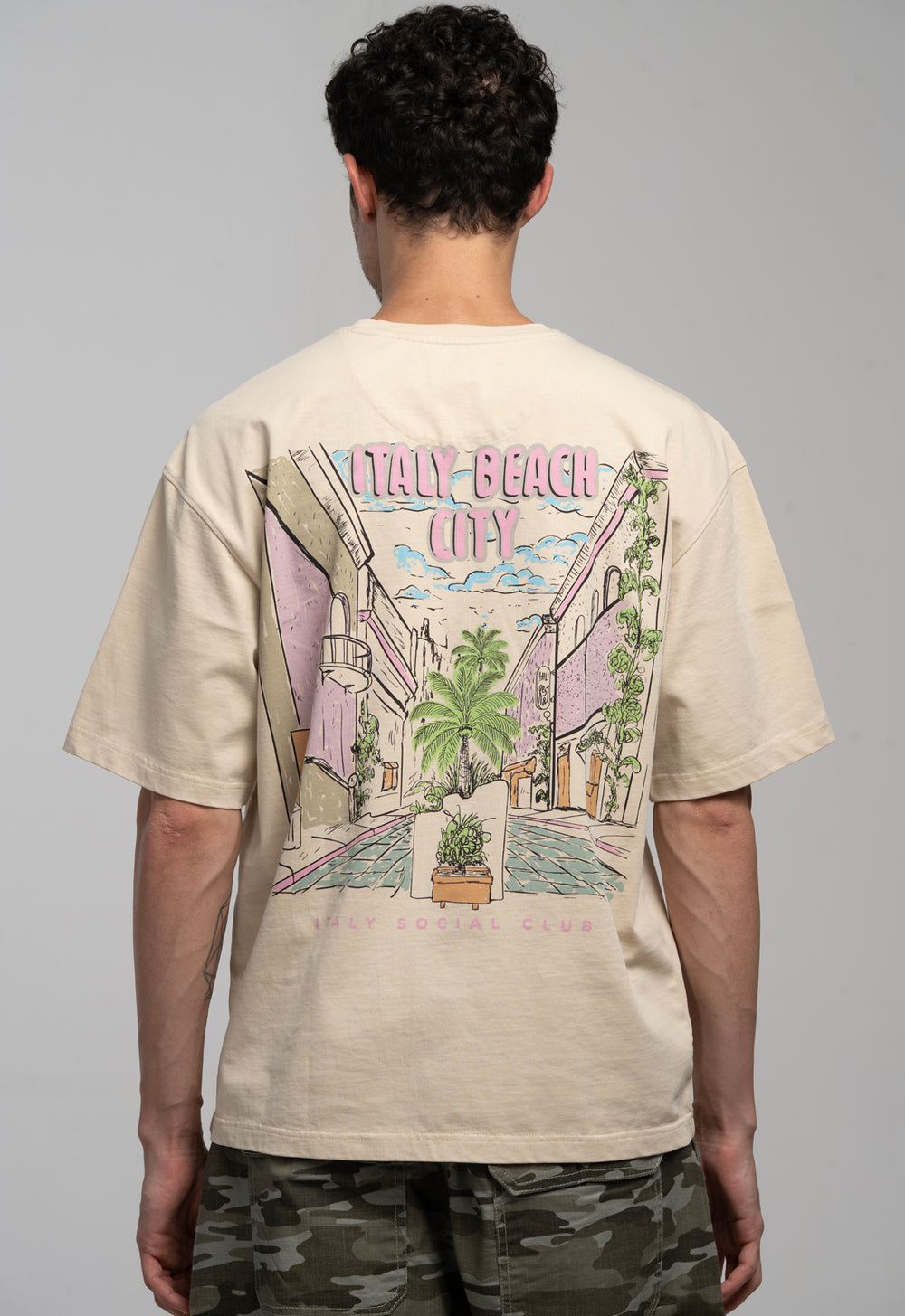 Person wearing a beige t-shirt with a colorful graphic on the back, standing against a plain background