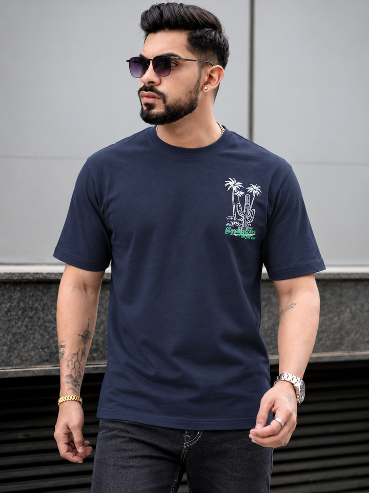 A man wearing a navy blue crewneck t-shirt with a small white and green palm tree graphic on the left chest, styled with dark sunglasses and dark jeans.