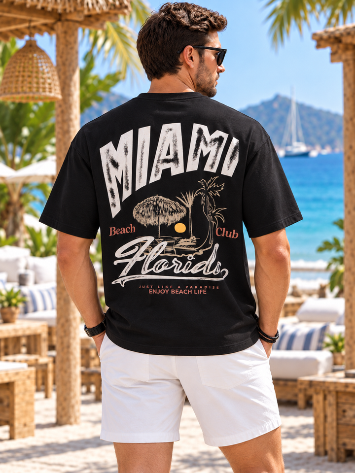 A man wearing a black graphic t-shirt with 'MIAMI Beach Club Florida' printed in white on the back, paired with white shorts, standing on a beach patio with a resort background.