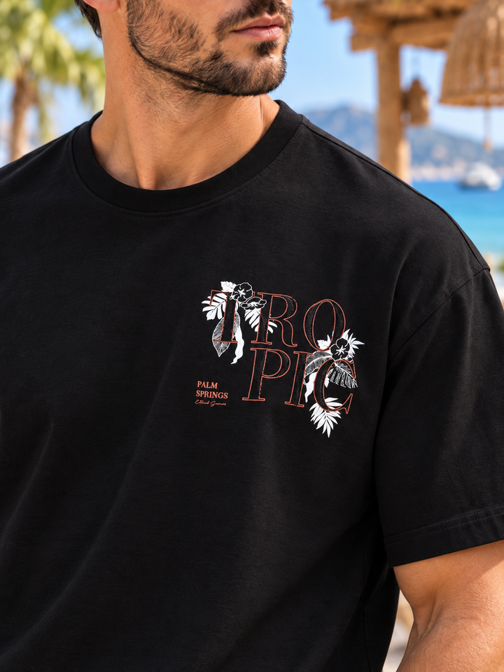 A man wearing a black short-sleeve t-shirt with a tropical graphic on the chest. The graphic features the word 'TROPIC' in a reddish-brown outline font, decorated with white palm tree illustrations, and includes the text 'PALM SPRINGS Florida' in a small red font.