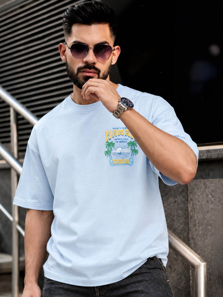 A man wearing a light blue crewneck t-shirt with a colorful 'Hawaii' graphic on the chest, styled with dark sunglasses, a watch, and dark jeans, standing in front of a modern building background.