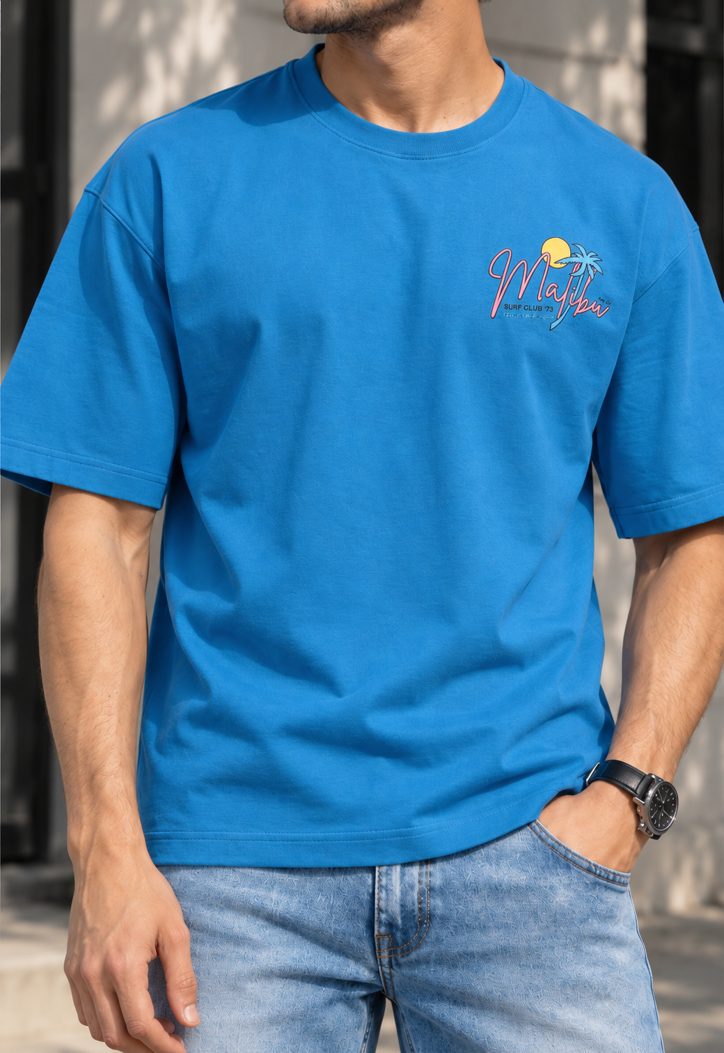 A man wearing a bright blue crewneck t-shirt with a small 'Maybo' graphic on the left chest, paired with light wash blue jeans.