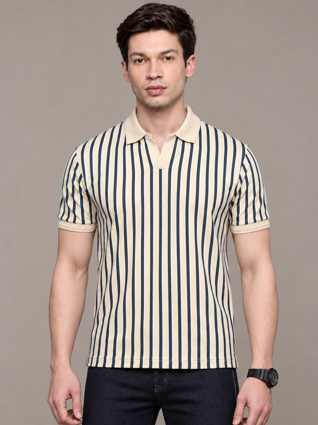 A man wearing a short-sleeve polo shirt with vertical navy blue and cream stripes, paired with dark jeans, standing against a solid gray studio background.