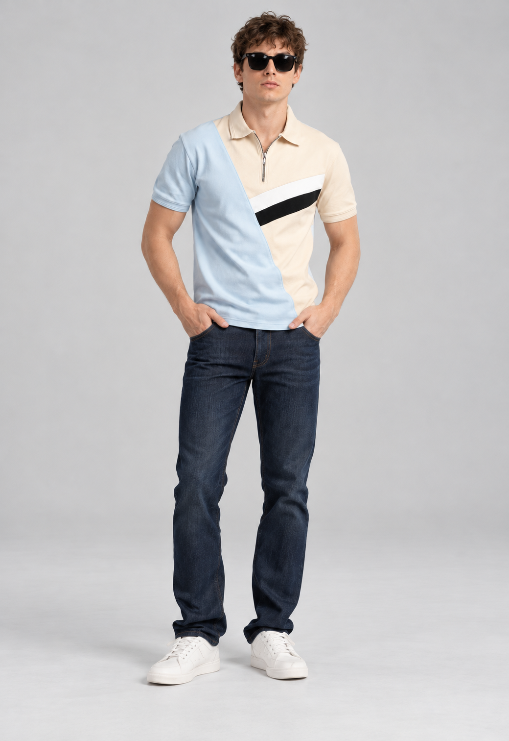 A man modeling a short-sleeve polo shirt with a diagonal color-blocked design in light blue, cream, and black, paired with dark blue jeans and white sneakers against a light gray studio background.