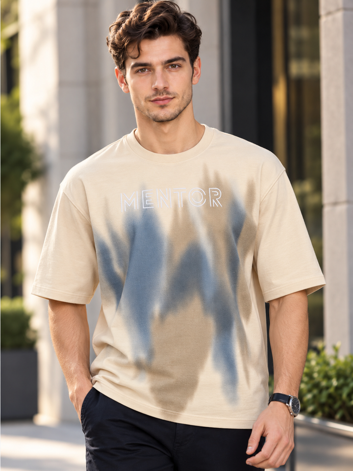 A man wearing a beige crewneck t-shirt with a blue and beige abstract graphic and the word 'MENTOR' printed across the chest, standing outdoors in front of a modern building.