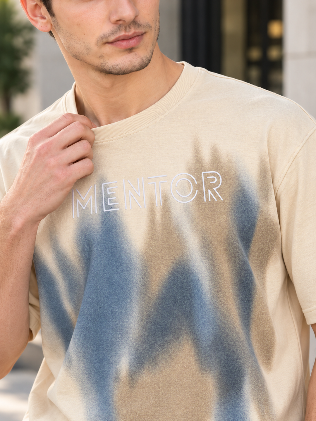 A close-up of a man wearing a beige crewneck t-shirt with a blue tie-dye splash pattern and the word 'MENTOR' outlined in white across the chest.
