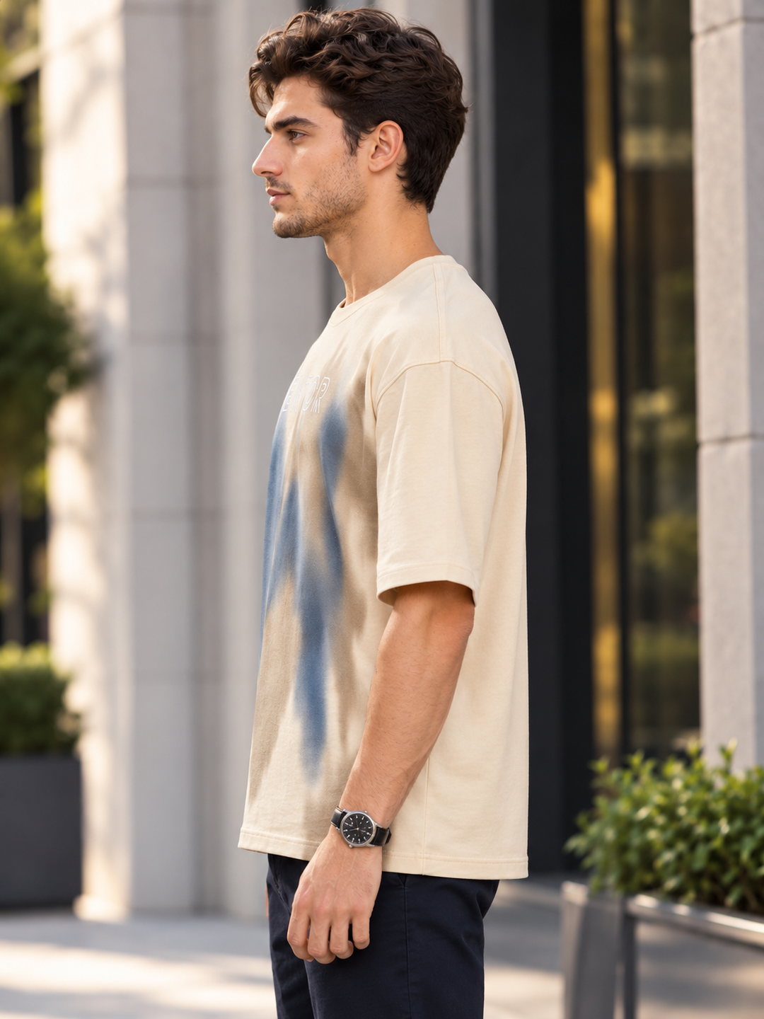 A man wearing a cream-colored short-sleeve t-shirt with a blue abstract graphic print, standing in an outdoor urban setting by mentor.